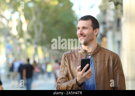 Front view portrait of a happy man holding smart phone walking towards camera looking at side in the street Stock Photo