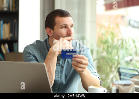 Pensive man holding a credit card thinking looking away through a window sitting in a coffee shop Stock Photo