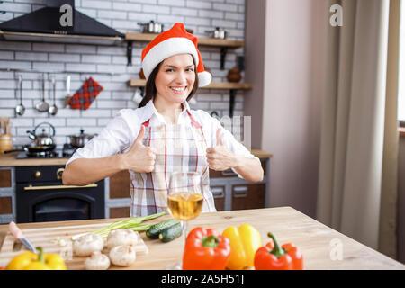 Festive attractive adult woman hold both big thumbs up and smile. Celebrating new year or Christmas. Cooking alone. Daylight. Looking on camera Stock Photo