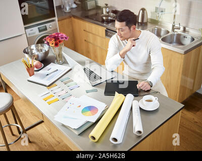 high angle view of a young asian designer sitting at kitchen counter working on a design using laptop computer and digital pen tablet Stock Photo