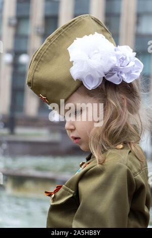 Little girl dressed in military uniform on background of fountains near the government building. White bow on her head. Children of war. Defenders of Stock Photo