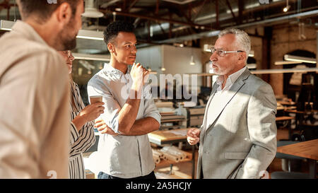 Discussing new project. Three young colleagues talking about something with confident mature man in formal wear while standing in the modern office Stock Photo