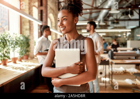 Modern business woman. Young and cheerful afro american woman holding laptop and smiling while standing in the modern office Stock Photo