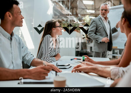 Brainstorming. Confident mature businessman using a whiteboard and explaining something to his young colleagues during meeting in the office Stock Photo