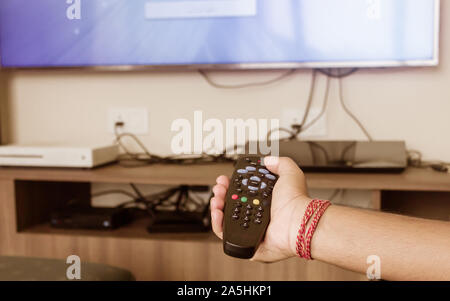 Close up of Human Hand holding a Remote control and pointed towards a TV screen for changing or zapping channels. Modern technology and lifestyle conc Stock Photo