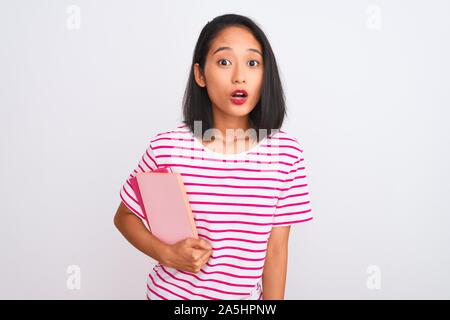 Young beautiful chinese woman holding books standing over isolated white background scared in shock with a surprise face, afraid and excited with fear Stock Photo