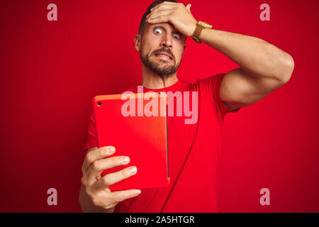 Young man using digital touchpad tablet over red isolated background stressed with hand on head, shocked with shame and surprise face, angry and frust Stock Photo