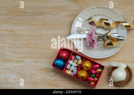 Beautiful Easter table settings decoration, white bunny, colorful chocolate eggs and plate with cutlery and spring flower on wooden background.Copy sp Stock Photo