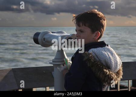Young child looking to side next to large scale public binoculars on Hastings pier in daytime Stock Photo