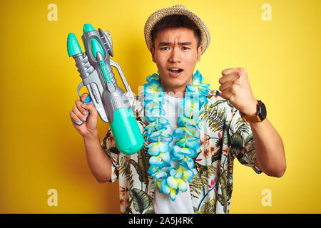 Asian chinese man wearing floral hawaian lei and water gun over isolated yellow background annoyed and frustrated shouting with anger, crazy and yelli Stock Photo