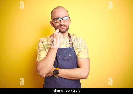Young bartender man wearing barista apron working as professional over yellow background with hand on chin thinking about question, pensive expression Stock Photo