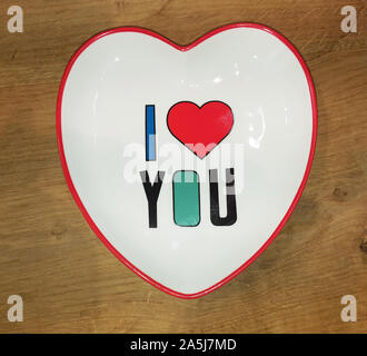 Ceramic white dish with a red edging, made in the shape of a heart with a multi-colored inscription  I LOVE YOU Stock Photo