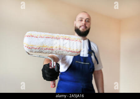 Construction worker at construction site with uniform, construction tool, equipment. He in blue coverall and holds paint roller brush for wall. Stock Photo