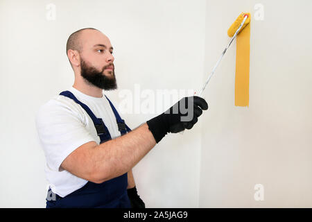 Construction worker doing finish renovation at apartment. Professional painter using paint roller brush painting of wall with yellow color. Stock Photo