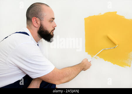 Construction worker doing finish renovation at apartment. Professional painter using paint roller brush painting of wall with yellow color. Stock Photo