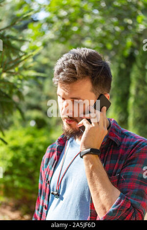 Handsome young brunette caucasian man tilted his head and speaks on the phone. Portrait of a man with a beard and mustache on a natural background, mo Stock Photo