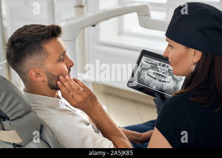 View from side of bearded man sitting on dental chair and keeping cheek with hand. Young patient with toothache listening female dentist and looking at x ray picture of jaw. Concept of medicine. Stock Photo
