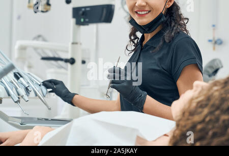 Cropped view of cheerful female dentist in uniform and protective gloves working with professional equipment in dental clinic. Woman smiling and curing teeth of patient. Concept of care. Stock Photo