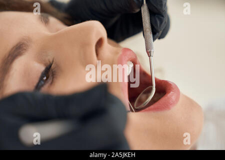 Closeup female face with opened mouth and hands of dentist keeping probe and mirror in clinic. Woman visiting dental office and curing teeth. Concept of treatment, care and health. Stock Photo