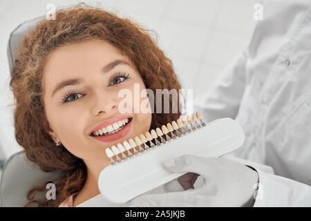 Closeup of beautiful young woman lying on dental chair, looking at camera and smiling while male dentist keeping in hands teeth color range. Girl doing whitening procedure in dental office. Stock Photo