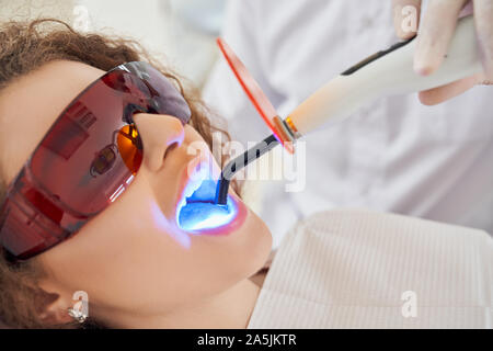 Closeup of young female patient lying on dental chair in protective gloves doing whitening procedure in clinic. Male dentist keeping ultraviolet tool. Concept of medicine and dentistry. Stock Photo