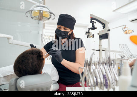 View from back of male patient visiting dental office. Professional female dentist in uniform and mask keeping probe and curing teeth. Special equipment and tools at background. Stock Photo