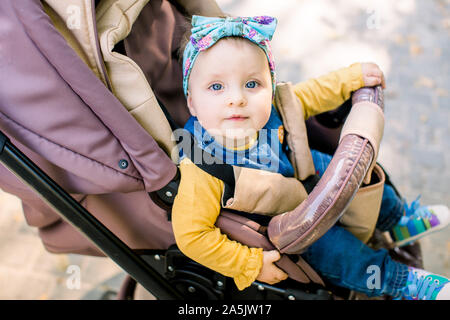 Happy girl in baby carriage playing in pram on background nature. Portrait cute little beautiful girl of 9 months sitting or stroller and waiting for Stock Photo
