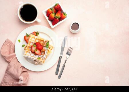 Belgian waffles with strawberries and cup of coffee on a pink background, table top view with copy space. Tasty sweet breakfast food