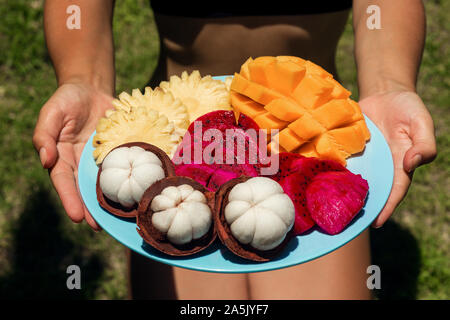 A girl in the green jungle is holding a plate with freshly cut and peeled tropical fruits. Stock Photo
