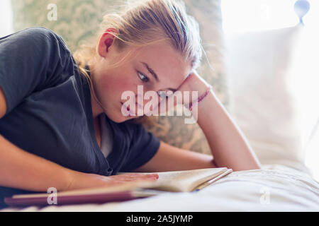 13 year old girl lying in her bed reading by window light Stock Photo