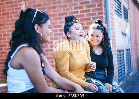 Young woman and her teenage sisters sitting on wall laughing Stock Photo