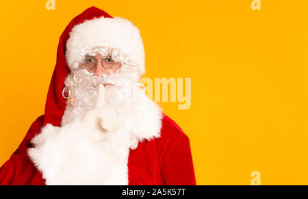 Santa Claus on orange background holding finger near his mouth Stock Photo