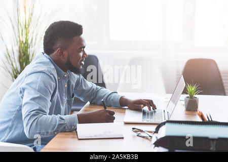 African worker taking notes while using laptop in office Stock Photo