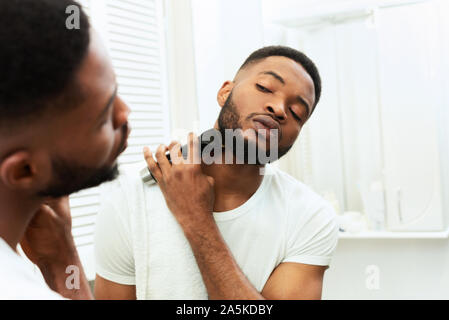Young black man looking at mirror and shaving beard with trimmer Stock Photo