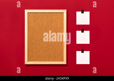 Empty cork board with wooden frame and three blank notes sticked with black tape on red background, top view with copyspace. Stock Photo