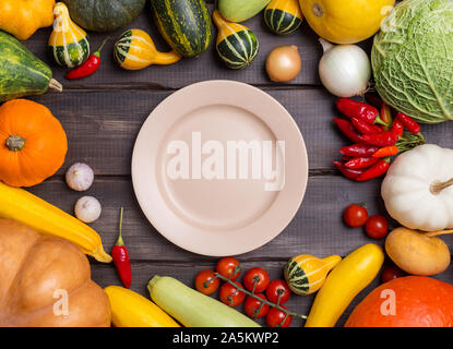 Empty plate with different kinds of harvest vegetables on dark wooden background. Pumpkin, cabbage, onion, tomatoes, garlic, pepper around the empty p Stock Photo