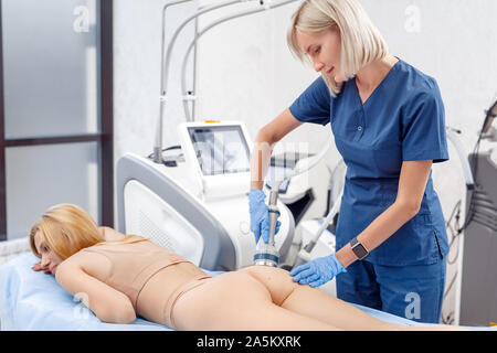 Cosmetology Service. Young woman at beauty clinic lying while doctor using body contouring coolwaves system tighten skin smiling joyful Stock Photo