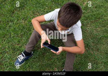 Close up of a little boy holding mobile phone in both hands while leaning arms on the knees. Concepts of dependence and education. Stock Photo