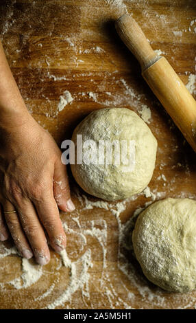 Preparing traditional homemade pasta. Fresh dough, rolling pin and woman's hand resting after kneading dough on floured wooden table. Top view Stock Photo