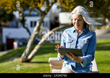 Smiling woman using tablet computer while standing by retaining wall in park during sunny day Stock Photo