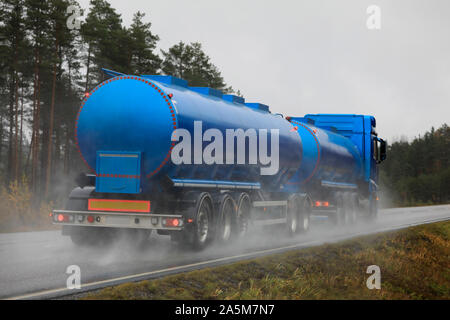 Blue tank truck for bulk transport hauls load on wet rural highway on a rainy day, rear view with water trail, Tenhola, Finland. October 18, 2019. Stock Photo