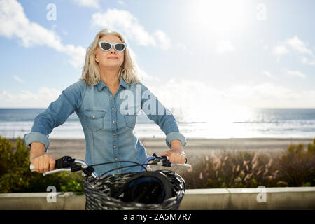 Senior woman wearing sunglasses riding bicycle against sea during sunny day Stock Photo