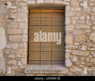 Closed window with wooden shutters and bars with a stone window sill in the old stone wall of the house Stock Photo
