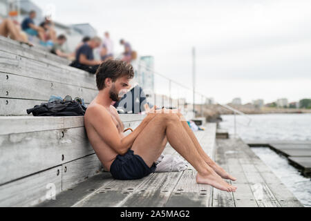 Man sitting on wooden steps and checking smart phone after swimming in the ocean Stock Photo