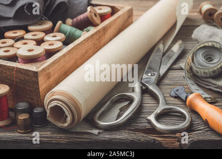 Sewing items: retro tailoring scissors, measuring tape, thimble, vintage spools of thread in wooden box, patterns on paper and tailor cutting knife. Stock Photo