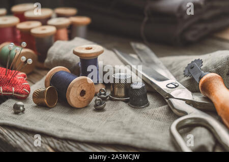 Retro sewing items: tailoring scissors, cutting knife, thimble, wooden thread spools, cushion for including pins, fabrics and sewing accessories. Stock Photo