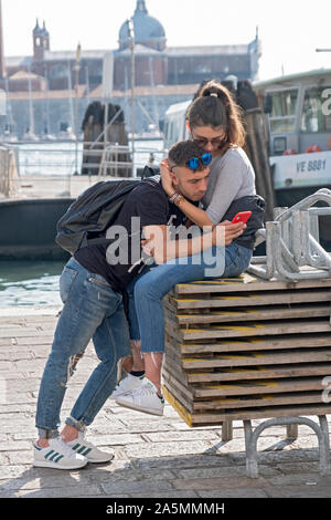 A young couple, likely tourists,  while the young man checks his cell phone. Near the Grand Canal in Venice, Italy. Stock Photo