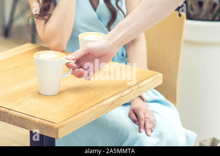 The male hand puts the cup of coffee on the table at cafe. Man hand is putting cup of coffee on the table on background of female in blue dress. Stock Photo