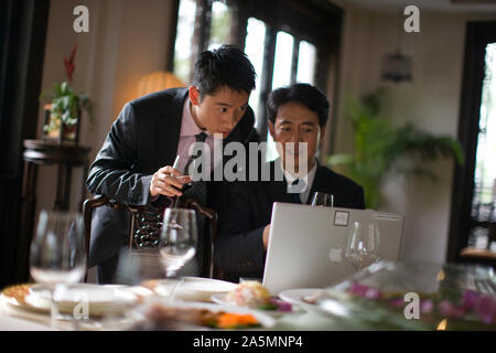 Two businessmen using a laptop while drinking wine at a table inside a restaurant. Stock Photo