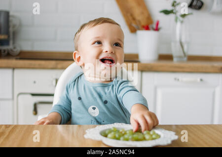 Charming little baby boy eating first food green grape at the bright kitchen at home Stock Photo